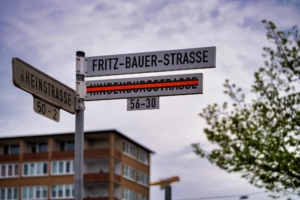 Foto vom neuen Straßenschild der Fritz-Bauer-Straße. Darunter hängt das mit leuchtend orange-farbenem Band durchgestrichene alte Schild der Hindenburgstraße.