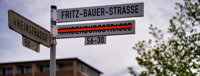 Foto vom neuen Straßenschild der Fritz-Bauer-Straße. Darunter hängt das mit leuchtend orange-farbenem Band durchgestrichene alte Schild der Hindenburgstraße.